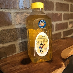 May include: A clear glass bottle of raw honey with a yellow lid. The label says "Outdoor Traditions Liquid Gold" and features a cartoon beekeeper with the words "Pure Raw Honey" underneath. The label also says "Made in Montana USA".