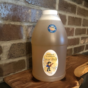 May include: A clear plastic bottle of honey with a yellow and blue label that says "Outdoor Traditions Liquid Gold Pure Raw Honey 1/2 US Gallon 2.25 Liters". The bottle is sitting on a wooden cutting board. The bottle has a white cap and a sticker that says "Made in Montana USA".