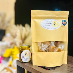 May include: A small glass jar of raw honey with a label that says "Raw Honey, Outdoor Tradition, Family Farm, Arizona" and a brown paper bag filled with honey caramels. The bag has a label that says "Honey Caramels, Homemade (small), Made with pure honey, cream, butter, brown sugar & vanilla."