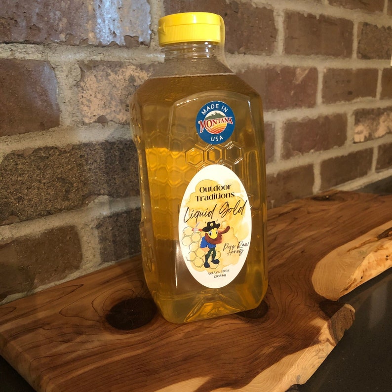 May include: A clear glass bottle of raw honey with a yellow lid. The label says "Outdoor Traditions Liquid Gold" and features a cartoon beekeeper with the words "Pure Raw Honey" underneath. The label also says "Made in Montana USA".