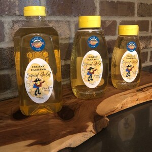 May include: Three glass bottles of honey with yellow lids. The bottles are labelled "Outdoor Traditions Liquid Gold Pure Raw Honey" and "Made in Montana USA".