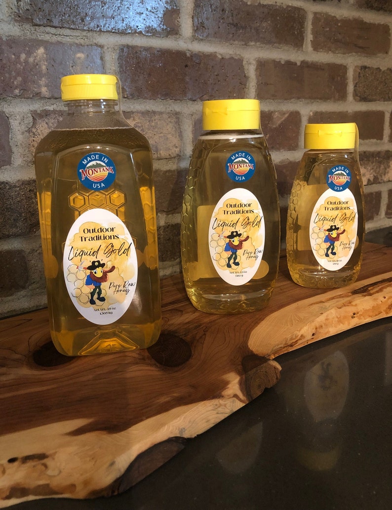May include: Three glass bottles of honey with yellow lids. The bottles are labelled "Outdoor Traditions Liquid Gold Pure Raw Honey" and "Made in Montana USA".