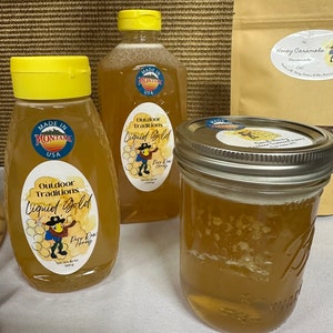 May include: Two glass jars of honey with a yellow label that reads "Outdoor Traditions Liquid Gold Pure Raw Honey". The jars are sitting on a white surface. A brown paper bag with the text "Honey Caramels Homemade (c)" is in the background.