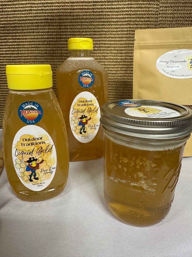 May include: Two glass jars of honey with a yellow label that reads "Outdoor Traditions Liquid Gold Pure Raw Honey". The jars are sitting on a white surface. A brown paper bag with the text "Honey Caramels Homemade (c)" is in the background.