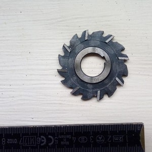 Puede incluir: Fresa circular de metal gris oscuro, con borde exterior dentado y orificio central. La fresa está sobre una superficie de madera clara. Una regla está en primer plano. Diámetro aproximado de 6 cm.