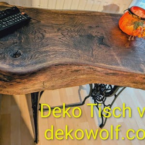 Könnte beinhalten: Ein rustikaler Holztisch mit Baumkante, getragen von einem schwarzen Metallgestell. Die Tischplatte ist braun mit sichtbarer Holzmaserung. Eine schwarze Fernbedienung und ein dekorativer Kürbis mit Kerze liegen auf dem Tisch.