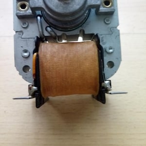 May include: A close-up shot of a small electric motor. The motor features a silver metal frame, a brown coil, and a central shaft. Two metal tabs extend from the base of the motor.