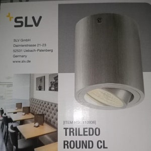 May include: A cylindrical, gray TRILEDO ROUND CL spotlight with an adjustable light source. The product packaging includes the SLV GmbH logo and company information. The image also shows a restaurant setting with tables and chairs.