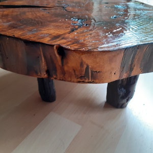 May include: A small, round wooden table with three dark brown legs. The tabletop is a natural wood slice with a glossy finish, showcasing the wood's grain and color variations. The table is approximately 12 inches in diameter.