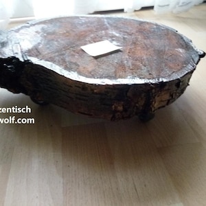 May include: A rustic, round wooden plant stand with a natural, bark-covered edge. The stand is brown with a lighter-colored top and has a small, rectangular tag on top. The text "Pflanztisch dekowolf.com" is visible on the side.