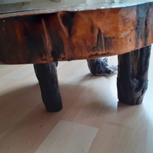 May include: A small, rustic wooden side table. The tabletop is a cross-section of a tree trunk, with a natural, varnished finish. The legs are dark, sturdy branches, giving it a natural, organic look.