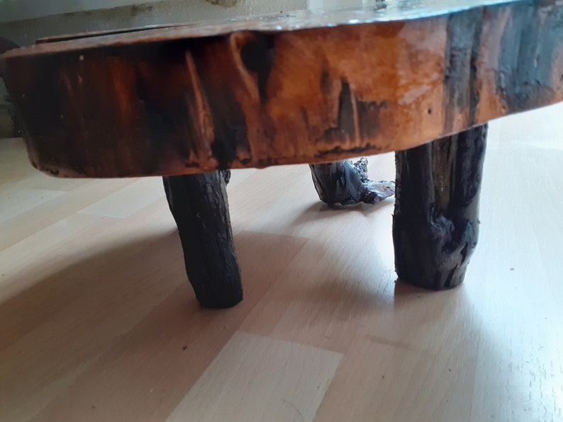 May include: A small, rustic wooden side table. The tabletop is a cross-section of a tree trunk, with a natural, varnished finish. The legs are dark, sturdy branches, giving it a natural, organic look.