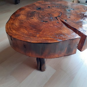 May include: A round, wooden coffee table made from a tree trunk slice. The table has a rich, dark brown color with visible wood grain and a glossy finish. It features a unique, organic shape and is supported by three small wooden legs.