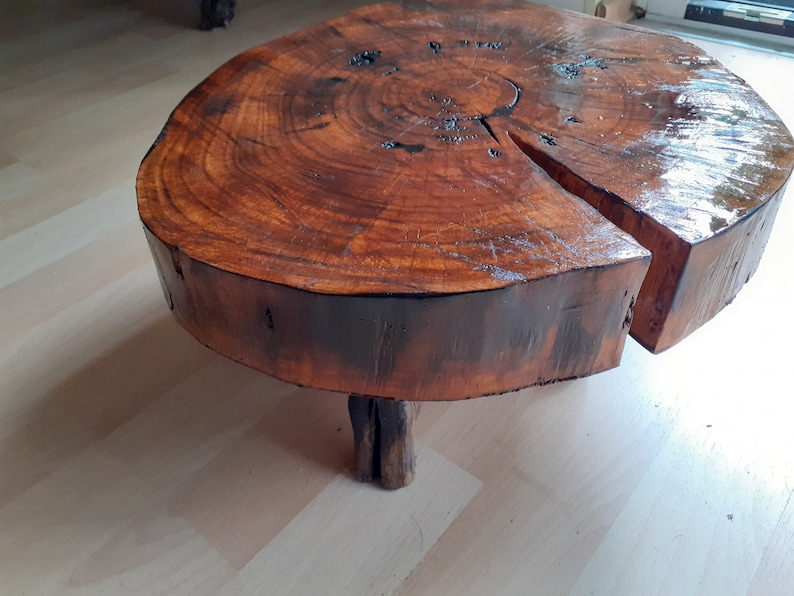 May include: A round, wooden coffee table made from a tree trunk slice. The table has a rich, dark brown color with visible wood grain and a glossy finish. It features a unique, organic shape and is supported by three small wooden legs.