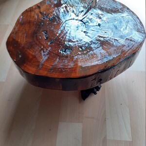 May include: A round, wooden side table with a natural, polished finish. The tabletop is a cross-section of a tree trunk, showcasing the wood's grain and color variations. The table is supported by a dark, decorative base.