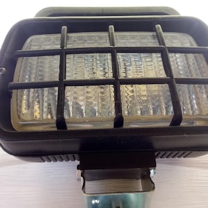 May include: A black rectangular work light with a protective metal grid over the glass lens. The light has a black rubberized housing and a metal mounting bracket. The lens is clear and textured.