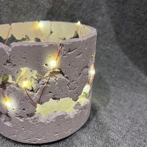 Può includere: Un vaso di cemento grigio chiaro con una superficie ruvida e testurizzata. Il vaso ha un'apertura circolare ed è decorato con una ghirlanda di luci di fata in filo di rame.