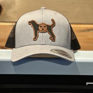 May include: A gray and black trucker hat with a brown leather patch featuring a black silhouette of a dog with a star on its back.
