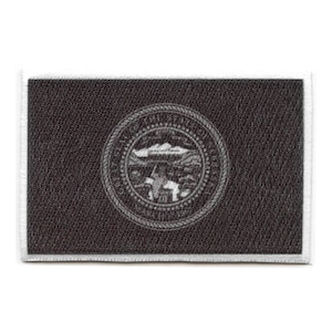 May include: Black and white embroidered patch featuring the state seal of North Dakota. The seal depicts a shield with a bison, a wheat stalk, and a sun rising over a mountain range. The words "State of North Dakota" are inscribed around the shield.
