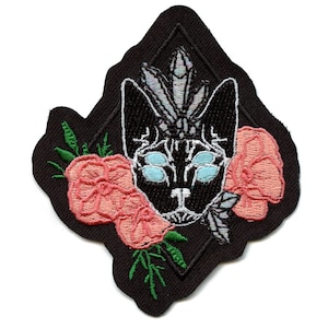 May include: Black and white embroidered patch of a cat's face with a crystal on its head, surrounded by pink flowers and green leaves on a black diamond-shaped background.