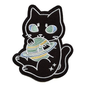 Op de afbeelding: Zwarte geborduurde strijkpatch van een kat die een groene planeet met een ring eromheen vasthoudt. De kat heeft blauwe ogen en een witte omtrek. Er zijn witte sterren op het lichaam van de kat.