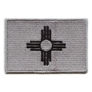 May include: A gray embroidered patch with the state symbol of New Mexico, a black Zia symbol, on a gray background.