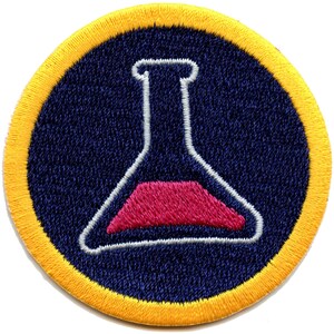 May include: A round, yellow embroidered patch with a dark blue center. The center features a white outline of a beaker with a pink liquid inside.