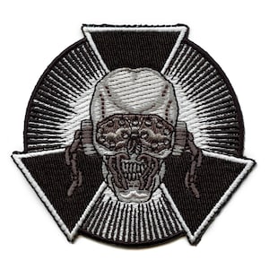 May include: Black and white embroidered patch featuring a skull with a cross behind it. The skull has a detailed design with a menacing expression. The cross is made up of three triangles.