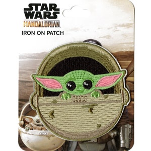 Op de afbeelding: Een opstrijkbare patch met het personage Grogu, ook bekend als Baby Yoda, uit The Mandalorian. De patch is groen met roze oren en staat tegen een beige achtergrond. De Star Wars- en The Mandalorian-logo's staan bovenaan.