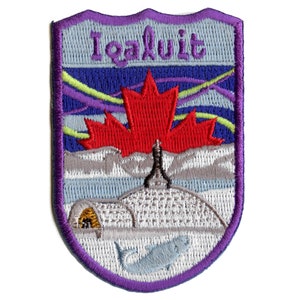 May include: An embroidered patch with a purple border, depicting a red maple leaf, a white igloo, a white fish, and the text "Iqaluit" in blue.