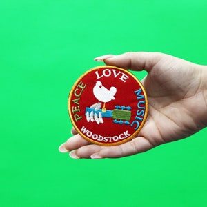 May include: A red, round embroidered patch with the words "Peace Love Music Woodstock" around a white dove holding a guitar.