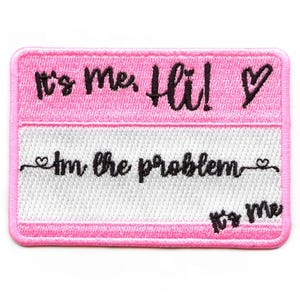 May include: A rectangular pink patch with the text "It's me, Hi!" above "I'm the problem" in black script. The bottom section is white with a black script that says "It's me". A black heart is next to "Hi!"