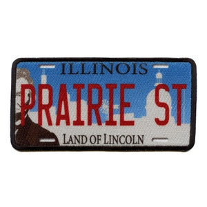 Op de afbeelding: Een blauw en wit Illinois kenteken met de tekst "PRAIRIE ST" in rode letters. De plaat heeft ook de tekst "ILLINOIS" en "LAND OF LINCOLN".
