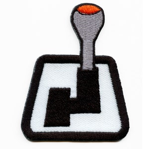 May include: A black and white embroidered patch of a stick shift gear lever with a gray shift knob and an orange top.