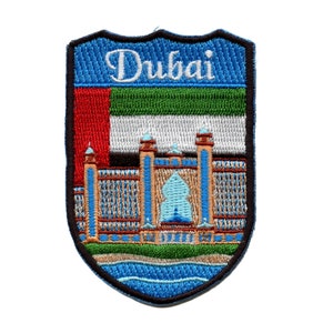 Op de afbeelding: Een geborduurde patch in blauw, groen, wit en rood met het woord "Dubai" in witte letters. De patch toont een gebouw met een grote boog en een blauw waterkenmerk. Het gebouw is omgeven door een groen gazon en een blauwe watermassa.