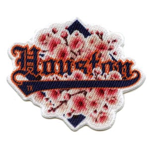 Può includere: Toppa ricamata con la scritta "Houston" in caratteri gotici arancioni e blu navy, su uno sfondo floreale di fiori rosa. La toppa ha un bordo bianco e le lettere "TX" sono in basso.