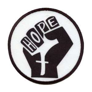 Pode incluir: Um remendo circular branco com um punho preto levantado e a palavra "HOPE" escrita nos nós dos dedos.