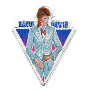 Può includere: Una toppa triangolare bianca con uno sfondo sfumato blu e viola. La toppa presenta una foto di David Bowie in un abito blu chiaro con una camicia bianca e una cravatta rossa. Il testo "DAVID BOWIE" è stampato in blu sulla toppa.