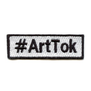 May include: A white embroidered patch with black stitching. The patch is rectangular and has a black border. The text "#ArtTok" is embroidered in black on the patch.