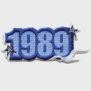 May include: Embroidered patch featuring the year "1989" in light and dark blue. The numbers have a textured appearance with a dark blue outline. Decorative star-like elements are on each side, with a white border.