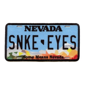 Könnte beinhalten: Ein blauer und weißer Nevada-Kennzeichen-Patch mit dem Text "SNAKE EYES" und dem Slogan "Home Means Nevada".