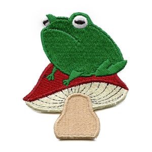 May include: Green frog sitting on a red and white mushroom with a tan stem. The frog has large eyes and a wide smile.