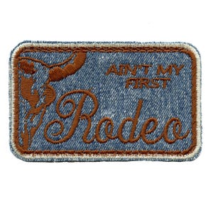 May include: A rectangular patch with a denim-like background and brown embroidered details. The patch features the words "AIN'T MY FIRST" above the word "Rodeo" in a decorative font. A brown silhouette of a rodeo rider is on the left.