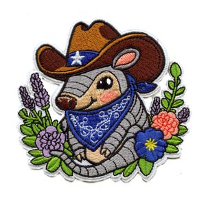 Puede incluir: Parche bordado con un armadillo de dibujos animados con un sombrero de vaquero y una bandana azul. El armadillo está rodeado de flores en tonos morados, rosas y azules, con hojas verdes. El sombrero de vaquero tiene una estrella blanca.