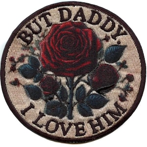 May include: Embroidered patch with a dark red rose and blue-green leaves. The patch has the text "BUT DADDY I LOVE HIM" in a brown, stylized font, set against a beige background with a brown border. The design is circular.