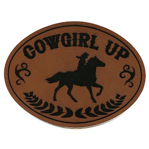 Peut inclure: Écusson ovale marron avec l'inscription "COWGIRL UP". Une silhouette noire d'une personne à cheval est au centre. Le design est encadré par des éléments décoratifs et une couronne de laurier.