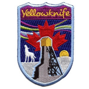 Peut inclure: Écusson brodé avec un fond bleu et un contour blanc. L'écusson présente une feuille d'érable rouge avec un bâtiment jaune et gris au centre. Le texte "Yellowknife" est en lettres jaunes sur un fond violet en haut de l'écusson.