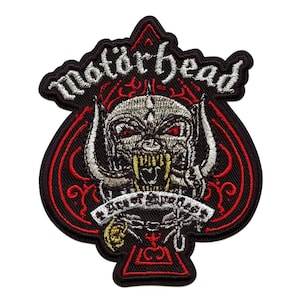 Peut inclure: Écusson brodé avec le nom du groupe "Motörhead" en écriture blanche. Il présente une illustration détaillée d'un visage démoniaque menaçant avec des yeux rouges et des dents jaunes. L'écusson est en forme de pique noire avec des détails rouges.