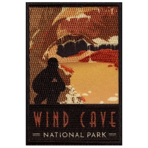 Op de afbeelding: Een geborduurde patch in zwart en bruin met een silhouet van een persoon die naar een grotingang kijkt. De tekst "WIND CAVE NATIONAL PARK" is onder de afbeelding gedrukt.