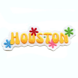 Può includere: Toppa ricamata con la scritta "Houston" in giallo, bordata di arancione, con accenti floreali colorati. La toppa è su sfondo bianco.